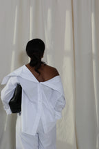 Woman wearing a pure cotton white off-shoulder button down shirt and white pants by Kureé The Brand Womenswear with a black leather bag, standing against a draped backdrop.