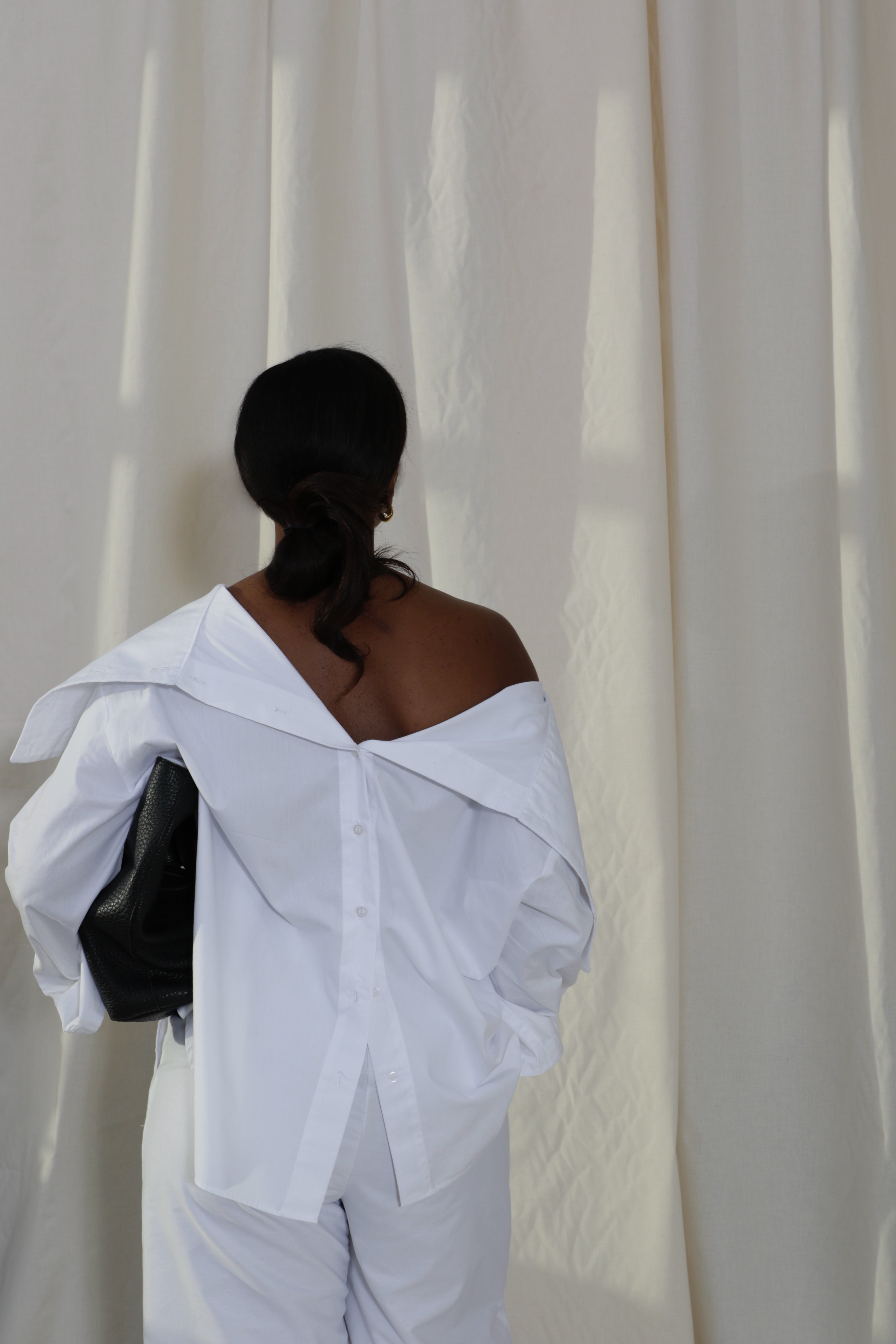 Woman wearing a pure cotton white off-shoulder button down shirt and white pants by Kureé The Brand Womenswear with a black leather bag, standing against a draped backdrop.