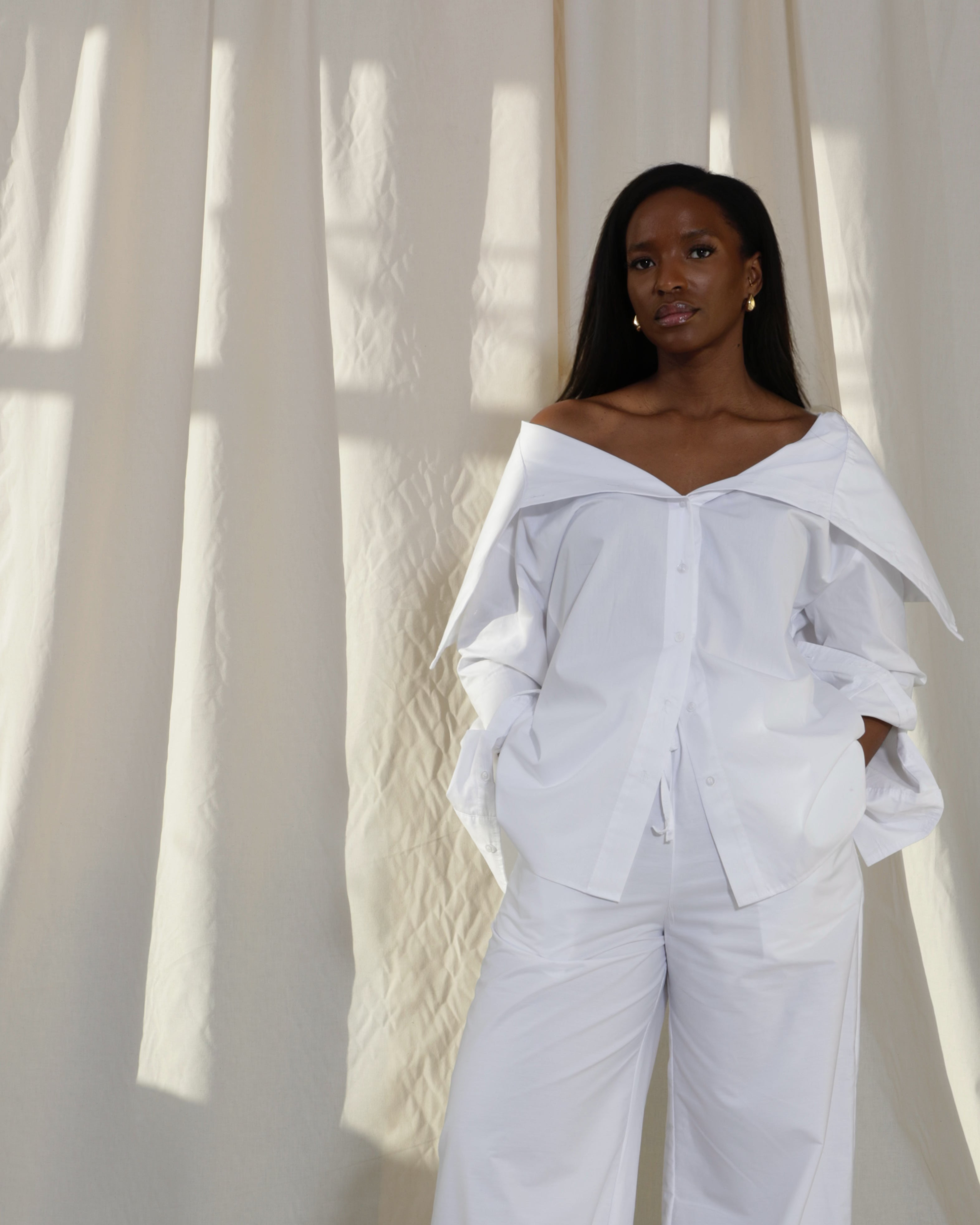 Woman wearing a pure cotton white off-shoulder top and white balloon trousers by Kureé The Brand Womenswear, standing against a draped backdrop.