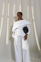 Woman wearing a pure cotton white off-shoulder top and white trousers by Kureé The Brand Womenswear with a black leather bag, standing against a draped backdrop.