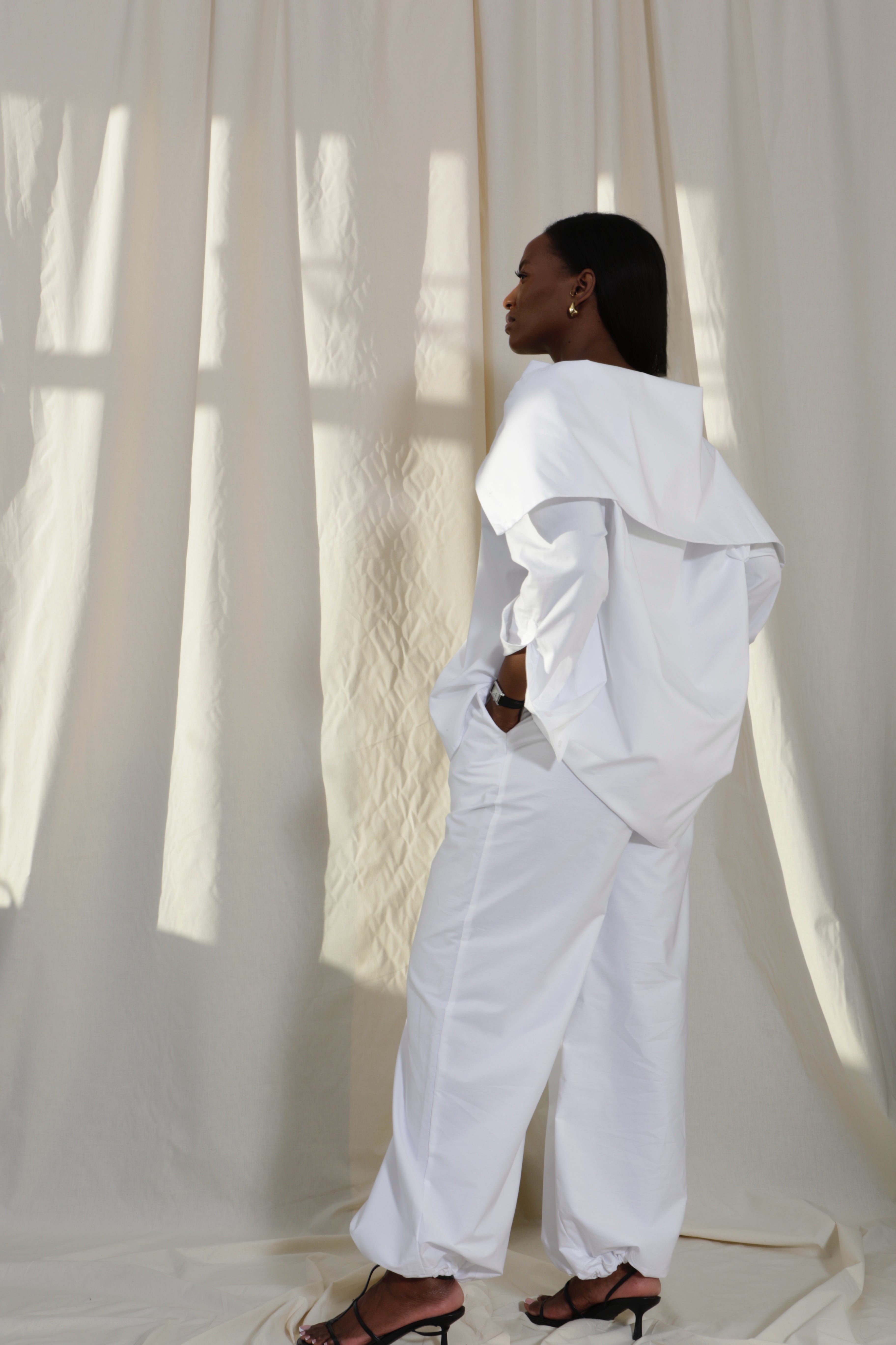 Woman wearing a pure cotton white off-shoulder top and white balloon trousers by Kureé The Brand Womenswear, standing against a draped backdrop.
